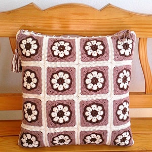 May include: A crocheted pillow with a brown and white floral pattern. The pillow is sitting on a wooden bench.