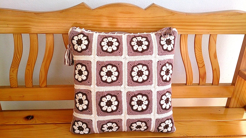 May include: A crocheted pillow with a brown and white floral pattern. The pillow is sitting on a wooden bench.