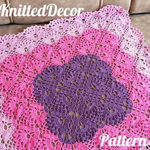 May include: A crocheted blanket with a square design in shades of pink and purple. The blanket has a scalloped edge and is made with a delicate stitch pattern. The text "Knitted Decor Pattern" is visible in the image.