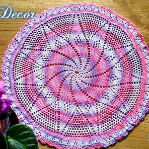 Puede incluir: Un tapete circular hecho a mano con un dise&ntilde;o en espiral en tonos rosa, blanco y morado. El tapete est&aacute; tejido a ganchillo con patrones intrincados y un borde festoneado. Las palabras "Knitted Decor" y "Pattern" son visibles.