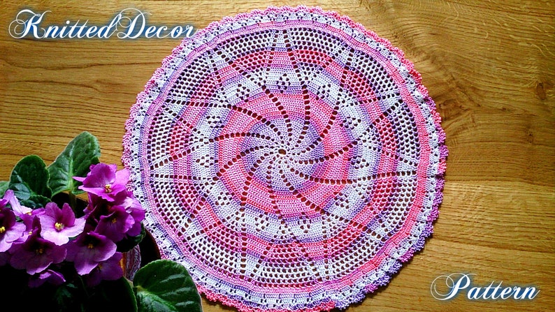 Puede incluir: Un tapete circular hecho a mano con un dise&ntilde;o en espiral en tonos rosa, blanco y morado. El tapete est&aacute; tejido a ganchillo con patrones intrincados y un borde festoneado. Las palabras "Knitted Decor" y "Pattern" son visibles.