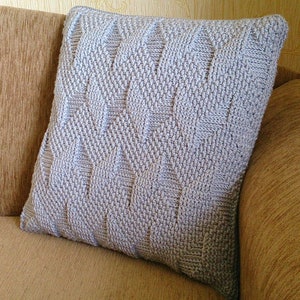 May include: A light grey, square cushion with a textured, woven design. The cushion is placed on a tan sofa. The design on the cushion features a repeating geometric pattern.
