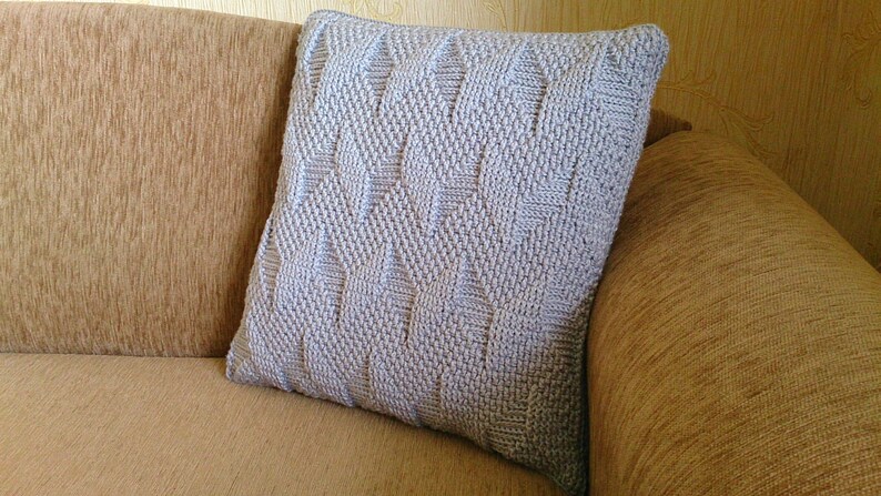 May include: A light grey, square cushion with a textured, woven design. The cushion is placed on a tan sofa. The design on the cushion features a repeating geometric pattern.