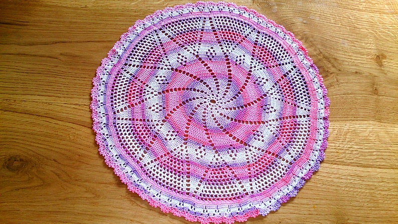 Puede incluir: Un tapete circular hecho a mano con un dise&ntilde;o en espiral. El tapete presenta una combinaci&oacute;n de hilos blancos, rosas y morados, creando un patr&oacute;n visualmente atractivo. Los bordes est&aacute;n acabados con un borde festoneado. Este art&iacute;culo decorativo es adecuado para la decoraci&oacute;n del hogar, aprox. 30 cm de di&aacute;metro.