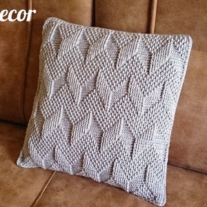 May include: A square, light grey knitted cushion with a textured diamond pattern. The cushion is on a brown sofa. The words "KnittedDecor" and "Pattern" are visible.