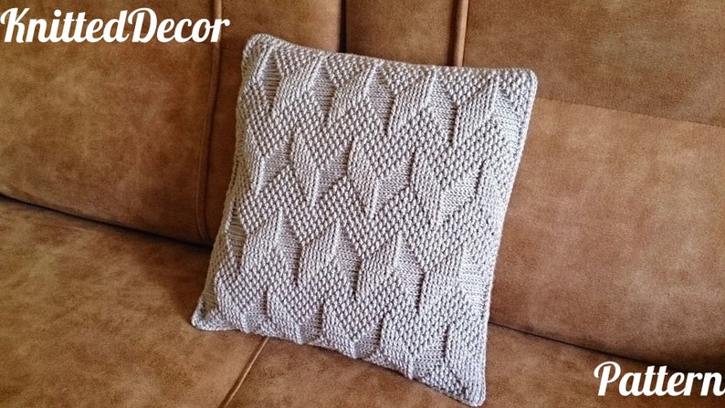 May include: A square, light grey knitted cushion with a textured diamond pattern. The cushion is on a brown sofa. The words "KnittedDecor" and "Pattern" are visible.