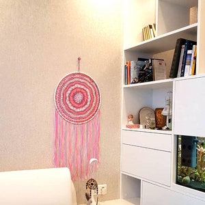 Puede incluir: Un atrapasue&ntilde;os rosa y blanco cuelga de una pared beige. El dise&ntilde;o circular presenta c&iacute;rculos conc&eacute;ntricos y largas hebras rosas y blancas. Un atrapasue&ntilde;os m&aacute;s peque&ntilde;o se encuentra sobre una mesita de noche blanca.