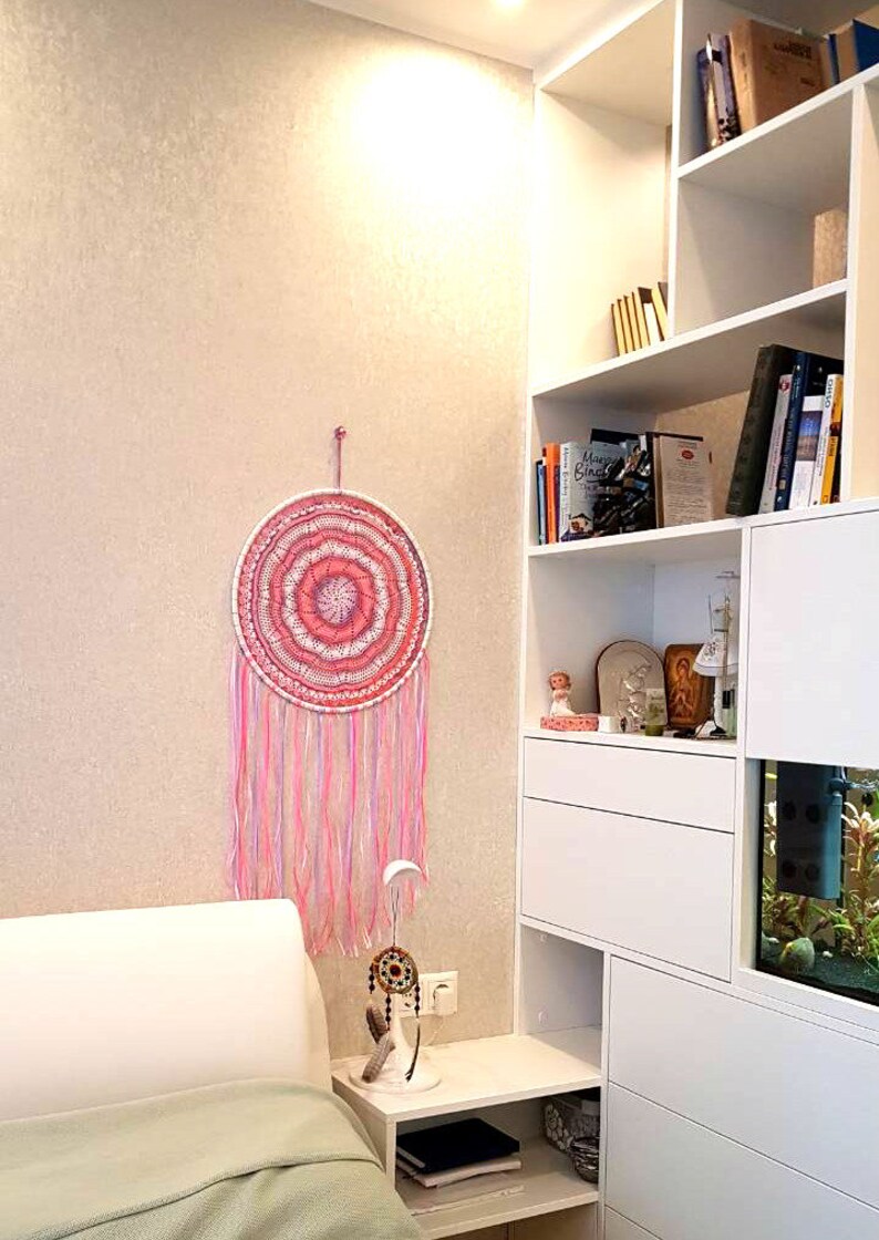 Puede incluir: Un atrapasue&ntilde;os rosa y blanco cuelga de una pared beige. El dise&ntilde;o circular presenta c&iacute;rculos conc&eacute;ntricos y largas hebras rosas y blancas. Un atrapasue&ntilde;os m&aacute;s peque&ntilde;o se encuentra sobre una mesita de noche blanca.