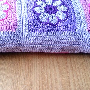 May include: A crocheted pillow with a purple, pink, and white floral pattern. The pillow is made of soft yarn and has a textured surface.