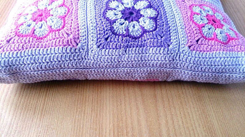 May include: A crocheted pillow with a purple, pink, and white floral pattern. The pillow is made of soft yarn and has a textured surface.