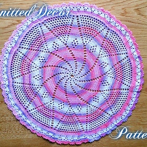 Puede incluir: Un tapete circular hecho a mano con un dise&ntilde;o en espiral. El tapete presenta un degradado de color rosa, blanco y morado, con un borde festoneado. Las palabras "Knitted Decor Pattern" est&aacute;n escritas en cursiva.