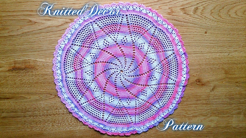Puede incluir: Un tapete circular hecho a mano con un dise&ntilde;o en espiral. El tapete presenta un degradado de color rosa, blanco y morado, con un borde festoneado. Las palabras "Knitted Decor Pattern" est&aacute;n escritas en cursiva.