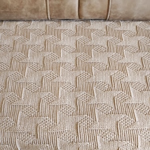 May include: A beige woven blanket with a geometric pattern of repeating shapes. The blanket is rectangular and appears to be made of a soft, textured material. The blanket is displayed on a brown sofa.