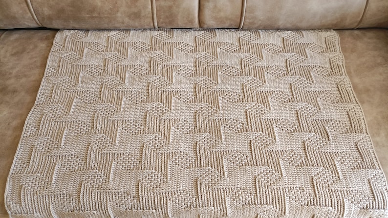 May include: A beige woven blanket with a geometric pattern of repeating shapes. The blanket is rectangular and appears to be made of a soft, textured material. The blanket is displayed on a brown sofa.