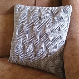 May include: A square, light grey knitted cushion with a geometric pattern of raised diamonds and rectangles. The cushion is on a brown sofa, a decorative accent for home decor.