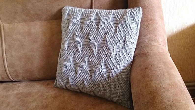 May include: A square, light grey knitted cushion with a geometric pattern of raised diamonds and rectangles. The cushion is on a brown sofa, a decorative accent for home decor.