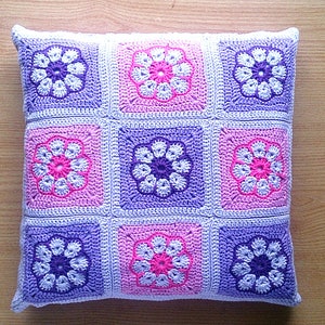 May include: A square crocheted pillow with a floral pattern in shades of pink, purple, and white. The pillow is made of soft yarn and has a textured surface.
