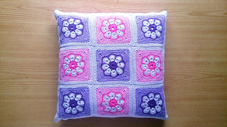 May include: A square crocheted pillow with a floral pattern in shades of pink, purple, and white. The pillow is made of soft yarn and has a textured surface.