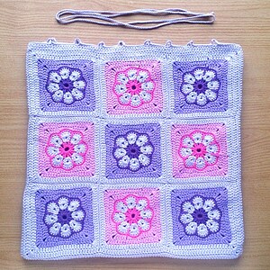 May include: A crocheted square blanket with a pattern of purple and pink flowers. The blanket is made of a soft, textured yarn and is perfect for adding a touch of handmade charm to any room.