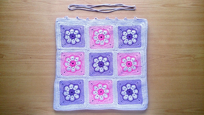 May include: A crocheted square blanket with a pattern of purple and pink flowers. The blanket is made of a soft, textured yarn and is perfect for adding a touch of handmade charm to any room.