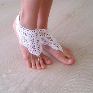 Könnte beinhalten: Weiße Häkel-Barfußsandalen. Diese zarten Sandalen haben ein florales Spitzendesign mit Zehenschlaufe und Knöchelriemen. Die Sandalen sind an den Füßen einer Person zu sehen, die auf einer hellen Holzmaserungsoberfläche ruhen.