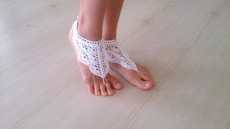 Pu&ograve; includere: Sandali a piedi nudi all'uncinetto bianchi. Questi delicati sandali presentano un design floreale in pizzo, con un anello per le dita e un cinturino alla caviglia. I sandali sono mostrati sui piedi di una persona, appoggiati su una superficie di legno chiaro.