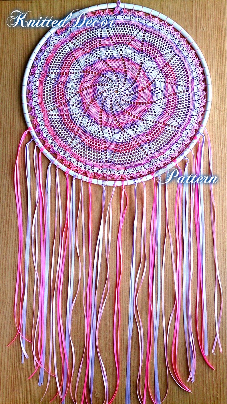 Puede incluir: Un atrapasue&ntilde;os hecho a mano con un tapete circular de ganchillo en tonos rosa, morado y blanco. El tapete est&aacute; colocado dentro de un aro blanco y adornado con largas cintas que fluyen en rosa, blanco y lavanda. Las palabras "Knitted Decor" y "Pattern" son visibles.