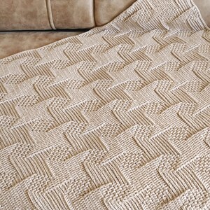 May include: A beige knitted blanket with a textured, geometric pattern. The blanket is folded over, displaying a repeating zig-zag design. The neutral color and detailed stitching suggest a handmade item, suitable for home decor.