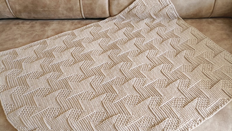 May include: A beige knitted blanket with a textured, geometric pattern. The blanket is folded over, displaying a repeating zig-zag design. The neutral color and detailed stitching suggest a handmade item, suitable for home decor.