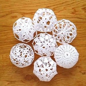 May include: Seven white, crocheted decorative spheres of varying designs are arranged on a wooden surface. The spheres have open, lace-like patterns, creating a delicate and airy appearance. The wooden surface has a warm, natural wood grain.