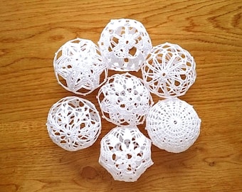 May include: Seven white, crocheted decorative spheres of varying designs are arranged on a wooden surface. The spheres have open, lace-like patterns, creating a delicate and airy appearance. The wooden surface has a warm, natural wood grain.