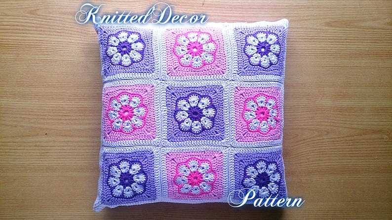 May include: A crocheted pillow with a floral pattern in shades of purple, pink, and white. The pillow is made up of nine squares, each with a different flower design. The text "Knitted Decor Pattern" is visible on the pillow.