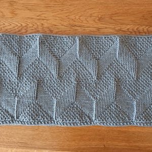 May include: A rectangular light blue knit scarf featuring a repeating geometric chevron pattern created with alternating textured knit and purl stitches. The fabric has a consistent light blue color throughout and a structured, dense knit construction.