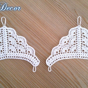 Pu&ograve; includere: Due pezzi decorativi bianchi all'uncinetto con bordo smerlato e un anello in alto per appendere. Il testo "Knitted Decor" &egrave; nell'angolo in alto a sinistra. Su una superficie di legno marrone chiaro.