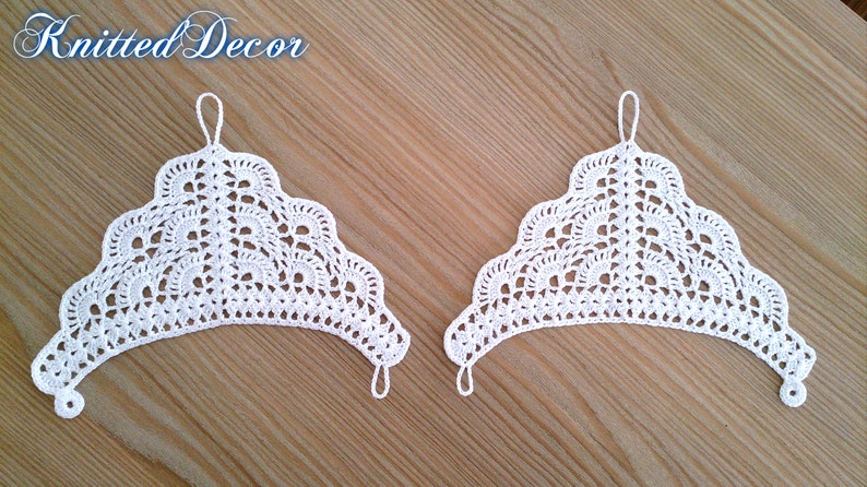 Pu&ograve; includere: Due pezzi decorativi bianchi all'uncinetto con bordo smerlato e un anello in alto per appendere. Il testo "Knitted Decor" &egrave; nell'angolo in alto a sinistra. Su una superficie di legno marrone chiaro.