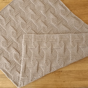 May include: Two light beige knitted blankets with a geometric pattern. One blanket is folded into a square, and the other is partially unfolded, revealing its rectangular shape. The blankets are made of a soft, textured material.
