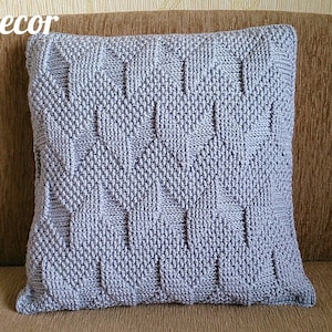 May include: A square, pale grey knitted cushion with a textured geometric design. The cushion is on a brown sofa. The words "Knitted Decor" and "Pattern" are visible.
