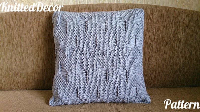 May include: A square, pale grey knitted cushion with a textured geometric design. The cushion is on a brown sofa. The words "Knitted Decor" and "Pattern" are visible.