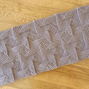 May include: A rectangular lavender knitted textile featuring a repeating geometric pattern of alternating vertical and horizontal textured stitches. The knit fabric displays a consistent light purple color throughout with a structured, woven appearance.