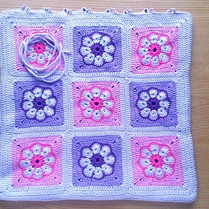 May include: A crocheted baby blanket with a floral pattern in shades of pink, purple, and white. The blanket is made up of nine squares, each with a different floral design.