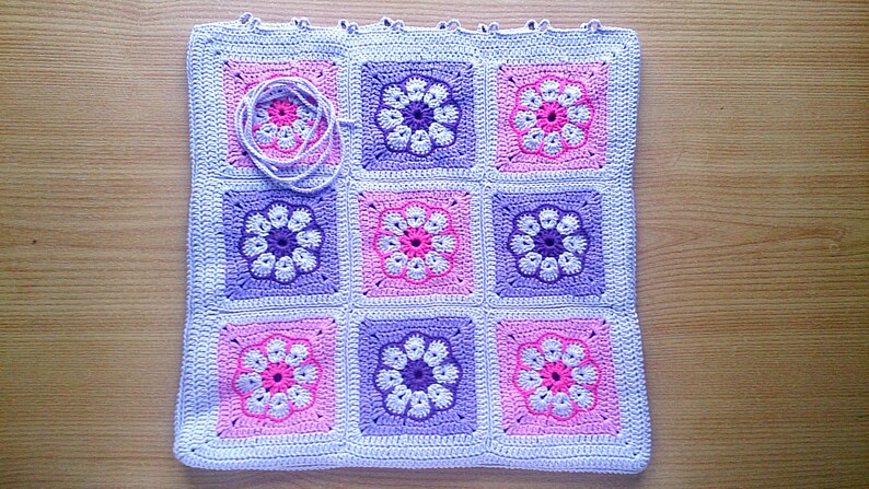 May include: A crocheted baby blanket with a floral pattern in shades of pink, purple, and white. The blanket is made up of nine squares, each with a different floral design.