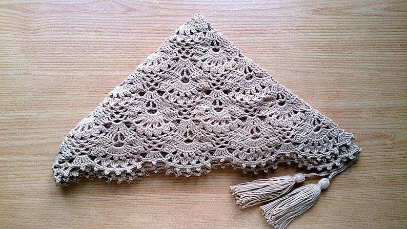 May include: A beige, triangular crochet shawl with a scalloped edge and two tassels. The shawl features a detailed, openwork pattern. The tassels are made of the same beige yarn as the shawl. The shawl is displayed on a wooden surface.