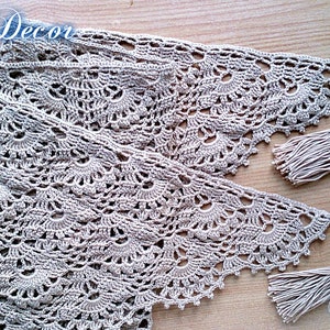 May include: A beige, knitted decor item with a delicate, openwork pattern. The item has a scalloped edge and is finished with tassels at each end. The text "Knitted Decor" and "Pattern" are visible in the image.