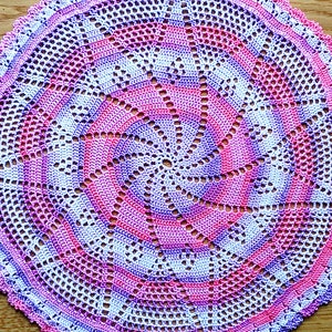 Puede incluir: Un tapete circular hecho a mano en tonos rosa, morado y blanco. El patr&oacute;n de ganchillo presenta un dise&ntilde;o en espiral en el centro, con un borde festoneado. El tapete est&aacute; colocado sobre una superficie de madera.