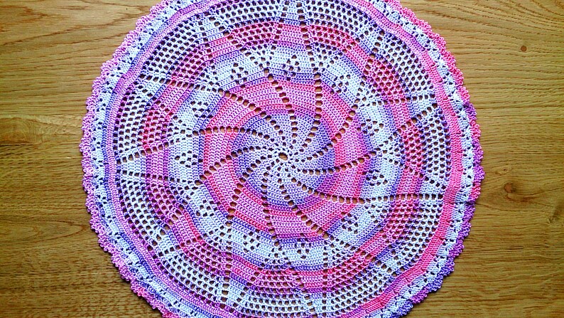 Puede incluir: Un tapete circular hecho a mano en tonos rosa, morado y blanco. El patr&oacute;n de ganchillo presenta un dise&ntilde;o en espiral en el centro, con un borde festoneado. El tapete est&aacute; colocado sobre una superficie de madera.