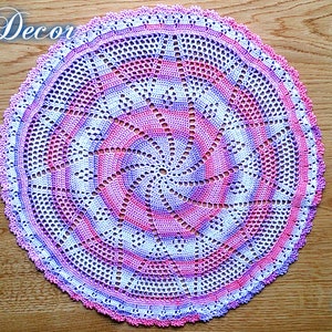 Puede incluir: Un tapete circular hecho a mano con un dise&ntilde;o en espiral en tonos de rosa, blanco y morado. El tapete est&aacute; tejido a ganchillo con intrincados calados y un borde festoneado. Las palabras "Knitted Decor" y "Pattern" est&aacute;n escritas en la imagen.