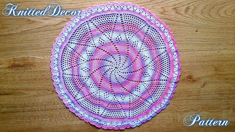 Puede incluir: Un tapete circular hecho a mano con un dise&ntilde;o en espiral en tonos de rosa, blanco y morado. El tapete est&aacute; tejido a ganchillo con intrincados calados y un borde festoneado. Las palabras "Knitted Decor" y "Pattern" est&aacute;n escritas en la imagen.