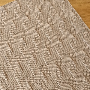 May include: A beige knitted blanket with a textured, geometric pattern. The design features a series of zigzags and diamond shapes, creating a visually interesting surface. The blanket is rectangular and appears to be made of soft yarn.