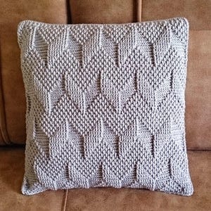 Pode incluir: Uma almofada decorativa quadrada cinza claro com um padrão geométrico. A almofada é feita de um tecido de malha texturizado e é colocada em um sofá de couro marrom. O design da almofada apresenta um padrão repetitivo de formas de diamante.