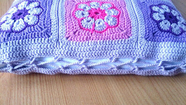 May include: A crocheted pillow with a purple, pink, and white floral design. The pillow has a white inner lining and a crocheted border.